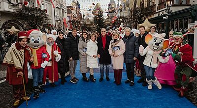 Die Inhaberfamilie Mack hat die Wintersaison im Europa-Park eröffnet. Foto: Europa-Park Foto von einer größeren Gruppe von Menschen, die auf einem blauen Teppich in einem Vergnügungspark stehen. Zwei der abgebildeten Personen sind als Mäuse verkleidet, drei weitere tragen ebenfalls Kostüme. Die anderen sind normal gekleidet.