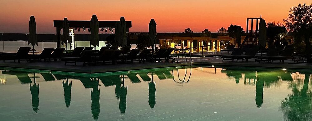 Luxushotel an der Ägäis: Wer früh bucht, sichert sich sein Wunschzimmer zu einem attraktiven Preis. Ob es der günstigte ist, weiß man erst später Ein ruhiger Außenpool bei Sonnenuntergang; das Wasser spiegelt die dunklen Umrisse von Liegen, geschlossenen Sonnenschirmen und einigen Bäumen. Am Horizont leuchtet ein sanfter Farbverlauf von Orange zu Dunkelblau, während rund um den Pool vereinzelte Lichter eine stimmungsvolle Atmosphäre erzeugen.