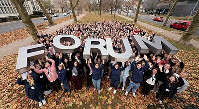 Rund 270 Reiseexperten diskutierten in Frankfurt am Main beim LCC-Forum über die Zukunft des Reisevertriebs Eine große Menschenmenge steht im Freien in einem herbstlichen Park und hebt gemeinsam ein großes, silberfarbenes 3D-Schriftobjekt mit dem Wort „FORUM“ in die Höhe. Die Personen lächeln, winken und wirken fröhlich; rundherum liegen gefallene Blätter, und im Hintergrund sind Bäume sowie angrenzende Straßen und Gebäude zu sehen.