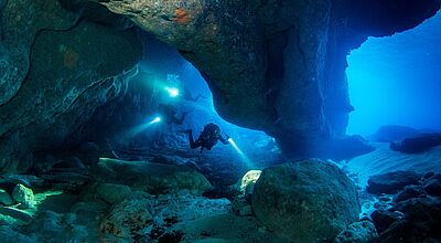 Rund um Malta können Höhlentaucher zum Beispiel Grotten oder Tunnel entdecken. Foto: Visit Malta Zwei Taucher unter Wasser, die mit einem Licht ausgestattet durch eine unterirdische Höhle schwimmen.