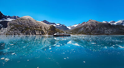 Holland America Line gilt als Pionier für Alaska-Kreuzfahrten. Foto: Holland America Line Ein Kreuzfahrtschiff mit dunklem Rumpf und weißem Aufbau passiert in Alaska gewaltige Gletscher.