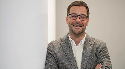 Neu an Bord bei der Schweizer Reederei: Ex Sea-Cloud-Cruises-Chef Daniel Schäfer Lächelnder Mann mit Brille in grauem Jackett und weißem Hemd, verschränkte Arme, an weißer Wand lehnend.