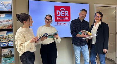 Auch Mike Brückner und sein Team vom Reisebüro Brückner sind jetzt DTPU-Partner Vier Mitarbeitende eines Reisebüros stehen vor einem Bildschirm mit dem Logo „DER Touristik Partner“ und blättern gemeinsam in Reisekatalogen. Im Hintergrund sind weitere DERTOUR-Kataloge im Regal zu sehen. Die Szene vermittelt Teamarbeit und Freude an der Beratung.