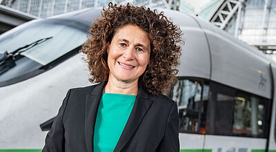Ist nur noch bis Jahresende bei der Bahn: Stefanie Berk, Marketing- und Vertriebschefin bei DB Fernverkehr Eine Managerin mittleren Alters mit dunklem Sakko und grüner Bluse steht lächelnd vor einem ICE-Zug vor einem Bahnhofsgebäude.