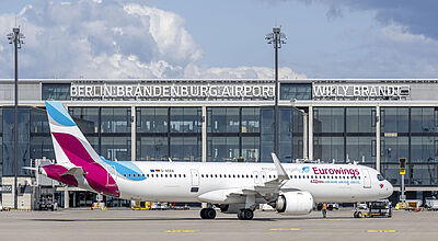 Mehr Flüge, mehr Maschinen: Eurowings investiert stark in den Standort Berlin Ein Flugzeug der Gesellschaft Eurowings steht vor dem Terminalgebäude des Flughafens Berlin Brandenburg an einem sonnigen Tag mit blauem Himmel und Wolken.