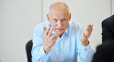 Klares Bekenntnis zum Reisebüro-Vertrieb: TUI-Chef Sebastian Ebel Ein Mann in hellblauem Hemd sitzt an einem Tisch, spricht gestikulierend mit erhobenen Händen und hat vor sich einige Dokumente liegen. Der Hintergrund ist schlicht und hell.